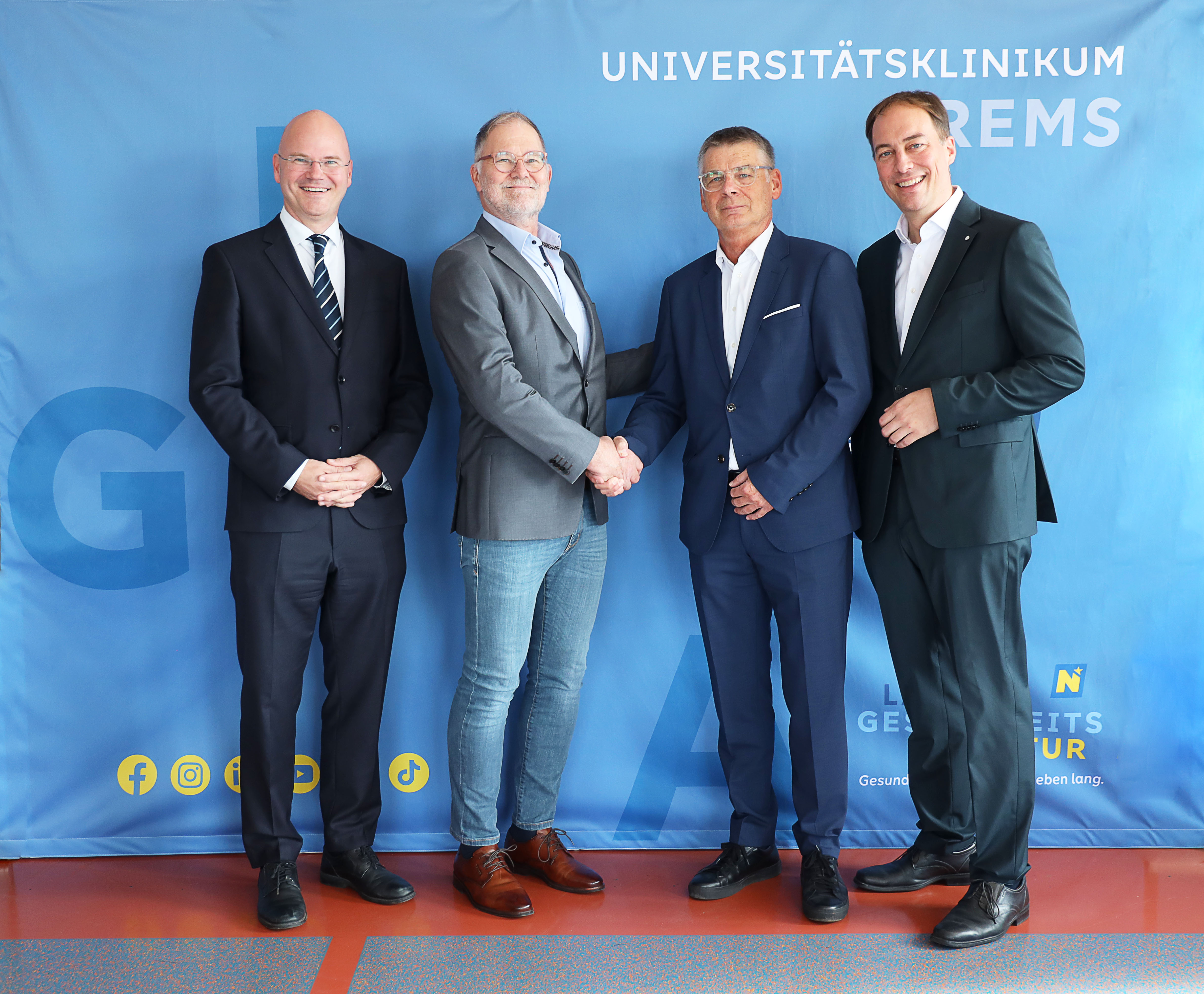 Universitätsklinikum Krems: Vier Männer in Anzügen und Jeans stehen vor blauem Banner, zwei schütteln Hände.
